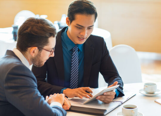 Closeup of two content adult business men sitting at table, using tablet computer, working with documents and discussing them in cafe