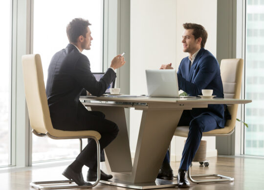 Two handsome businessmen sitting in comfortable chairs at desk with laptops in meeting room. CEO making important negotiation about companies partnership or corporate merger. Financiers planning deal